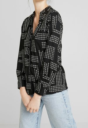 Kvinde iført en sort bluse med hvide pilmønstre og lyseblå jeans, stående med hænderne foldet foran sig mod en ensfarvet baggrund.