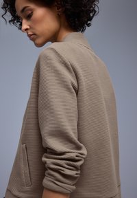 Jeune personne aux cheveux bouclés portant une veste beige texturée aux manches retroussées, regardant vers le bas sur un fond gris uni.