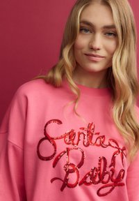 Rožinė palaidinė laisvu kirpimu, kuriame yra blizgių raudonų šilkinių raidžių užrašas "Santa Baby" ryškiu kursyvu priešakinėje pusėje. Lygaus audinio tekstūra.