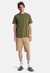 Olive green short-sleeve t-shirt with a textured finish, paired with beige shorts, white socks with logo, and dark green slip-on shoes.