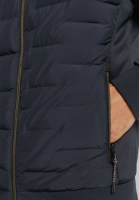 Pufferjack in donker marineblauw, met quilted ontwerp, ritssluiting met gouden details, zijzak en ribgebreide manchetten. Zachte textuur.