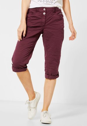 Mujer con pantalones capri burdeos remangados y zapatillas blancas, de pie frente a un fondo liso y claro.
