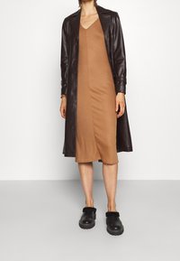 Abito marrone a coste con scollo a V, indossato sotto un lungo trench in pelle marrone scuro. Le scarpe foderate di pelliccia nera completano il look.