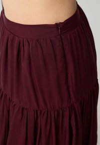 Bordeaux Maxi-Rock aus plissiertem Stoff, mit elastischem Bund und seitlichem Reißverschluss; glatte Textur und fließende Silhouette.