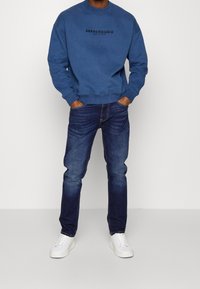 Blå bomullströja med svart logotyp, kombinerad med mörka denimjeans och vita sneakers. Tröjan har rundad hals och ribbade ärmslut.