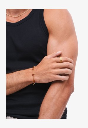 Uomo con canottiera nera che indossa una collana d'oro, un braccialetto d'oro con pietre rosse e un anello d'oro che si tiene il braccio superiore opposto.