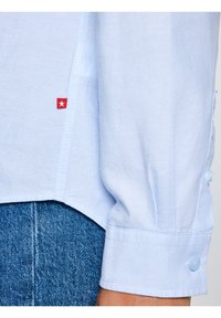 Gaiši zila krekla aproce aizpogāta pie plaukstas, valkāta virs zilām džinsa biksēm, ar nelielu sarkanu birku, kurā attēlota balta zvaigzne, pie krekla sānu šuves.