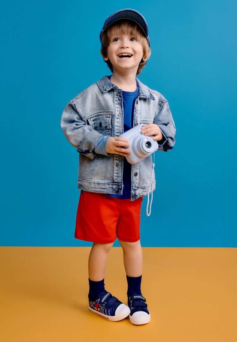 Jeune garçon en veste en jean, short rouge et casquette souriant tout en tenant un appareil photo instantané blanc sur un fond bleu et jaune.