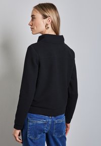 Giacca nera trapuntata con colletto alto, tessuto testurizzato e polsini a costine, abbinata a jeans blu in denim con tasche posteriori.