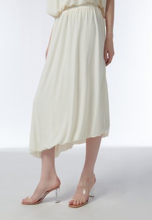 Vrouw draagt een crèmekleurige asymmetrische midirok en hooggehakte sandalen met doorzichtige bandjes op een effen witte achtergrond.