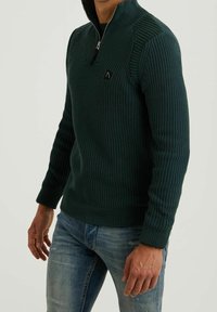 Zelený žebrovaný svetr s vysokým límcem, zapínáním na zip a malým logem. V kombinaci s světle modrými džínami s odřeným vzhledem.