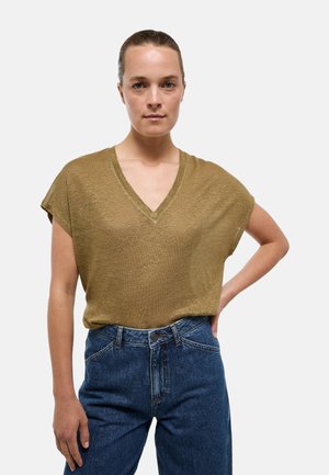 Femme debout, main sur la hanche, portant un haut marron à manches courtes et col en V, ainsi qu’un jean bleu taille haute sur un fond uni.