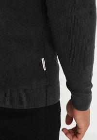 Gerippter dunkelgrauer Pullover mit langen Ärmeln, einem strukturierten Muster und einem kleinen Markenlabel am seitlichen Saum.