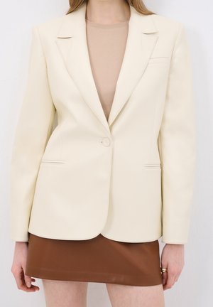 Woman wearing a cream single-button blazer over a beige top and a brown leather skirt, with hands relaxed at sides.