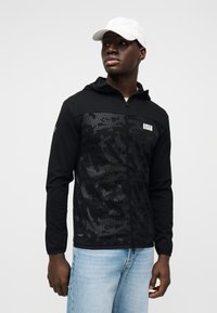 Μαύρο μπουφάν με φερμουάρ, διαθέτει υφή καμουφλάζ, ελαφρύ ύφασμα και κουκούλα. Συνδυάζεται με ανοιχτόχρωμα μπλε τζιν και λευκό καπέλο.