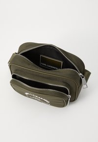 Borsa a tracolla verde oliva con materiale texture. Presenta due compartimenti con zip, hardware in argento e logo del marchio in bianco. L'interno è nero.