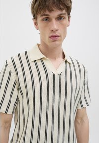 Kortærmet, cremet polo-shirt med sorte lodrette striber, et struktureret strikkemønster og en V-halskrave.