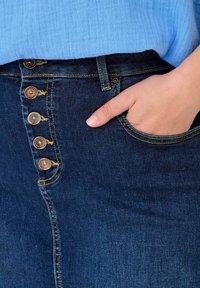 Temno modri jeans z visokim pasom, petimi bronastimi gumbi, kontrastnim rumenim šivom in roko v žepu.