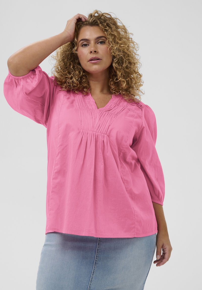 Blouse rose ample avec un décolleté en V, des détails plissés à l'avant et des manches trois-quarts. Le tissu semble léger et doux.