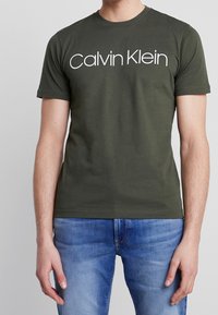 Olivově zelené bavlněné tričko s krátkými rukávy, s bílým logem "Calvin Klein" na hrudi. Volný střih, hladký materiál.