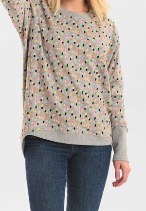 Kobieta w szarej bluzie z kolorowym wzorem abstrakcyjnych kropek i niebieskich dżinsach, podnosząca jedną rękę, kadrowana poniżej ramion.