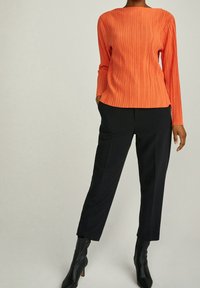 Top plissettato arancione a maniche lunghe abbinato a pantaloni neri su misura e stivaletti neri alla caviglia, che mettono in evidenza una silhouette elegante e una texture moderna.