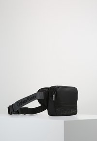 Schwarze Nylon-Tasche in rechteckiger Form, mit Reißverschluss, verstellbarem Gurt und geprägtem Logo auf der Vorderseite.