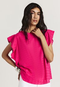 Blusa rosa in tessuto intrecciato con maniche corte a volant, caratterizzata da un motivo a diamante testurizzato e una vestibilità ampia, abbinata a pantaloni bianchi.