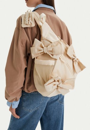 Mujer con vaqueros azules y chaqueta marrón lleva una mochila beige adornada con grandes lazos de tela y una pequeña bolsa adjunta.