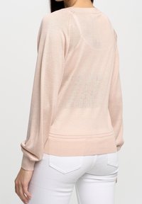 Helles rosa Strickpullover mit langen Ärmeln und einem gerippten Saum. Strukturierte Rückendetails und subtile durchsichtige Bereiche. Kombiniert mit weißen Jeans.