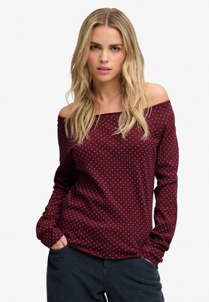 Kvinde med langt blond hår iført en burgundy off-shoulder top med polkaprikker og mørke bukser, hænder i lommerne, kigger fremad.