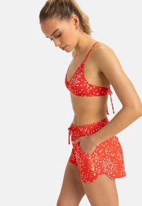 Röd bikini med vitt blommönster, som har en triangulär topp och matchande shorts med elastisk midja och sidofickor.