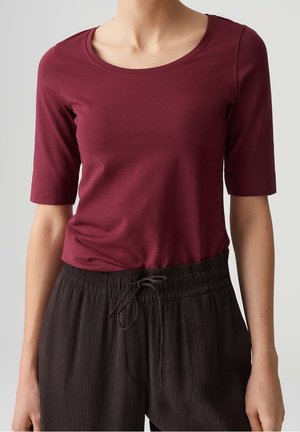 Femme portant un haut ajusté bordeaux à encolure dégagée avec des manches arrivant aux coudes, et un pantalon noir texturé ample avec cordon de serrage.