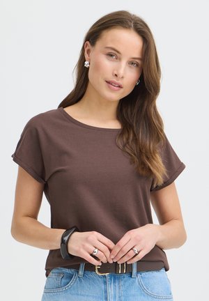 Ung kvinde med langt brunt hår iført en brun skjorte med op-rullede ærmer, blå jeans, sølvøreringe, ringe og et sort armbånd mod ensfarvet baggrund.