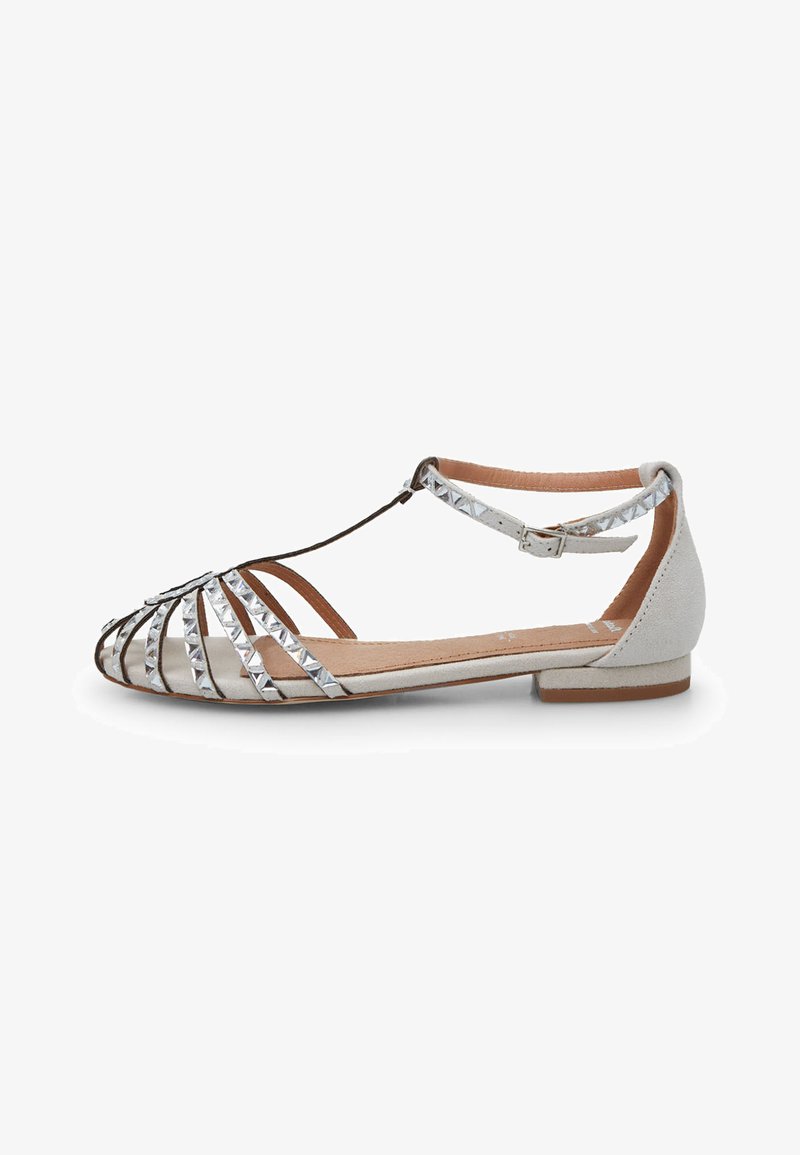 Flat sandal with grey suede material, silver pyramid studs, open toe, ankle strap, and a low heel, featuring a delicate crisscross design.