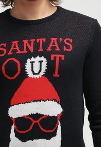 Schwarzer Strickpullover mit einem Santa-Gesicht, das einen roten Hut und Sonnenbrille trägt. Der Text lautet "SANTAS OUT" in fetten roten Buchstaben.
