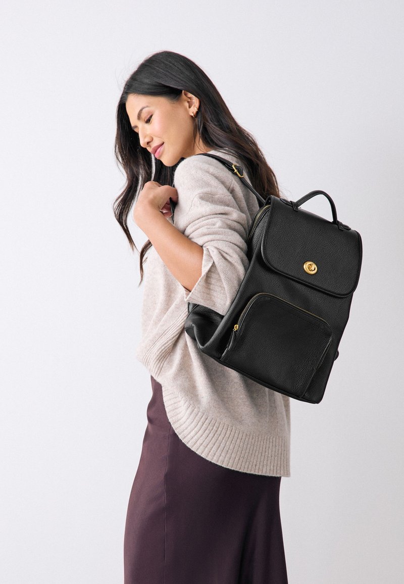 Frau mit langen dunklen Haaren, die einen beigen Pullover und einen lila Rock trägt, trägt einen schwarzen Leder-Rucksack über einer Schulter.