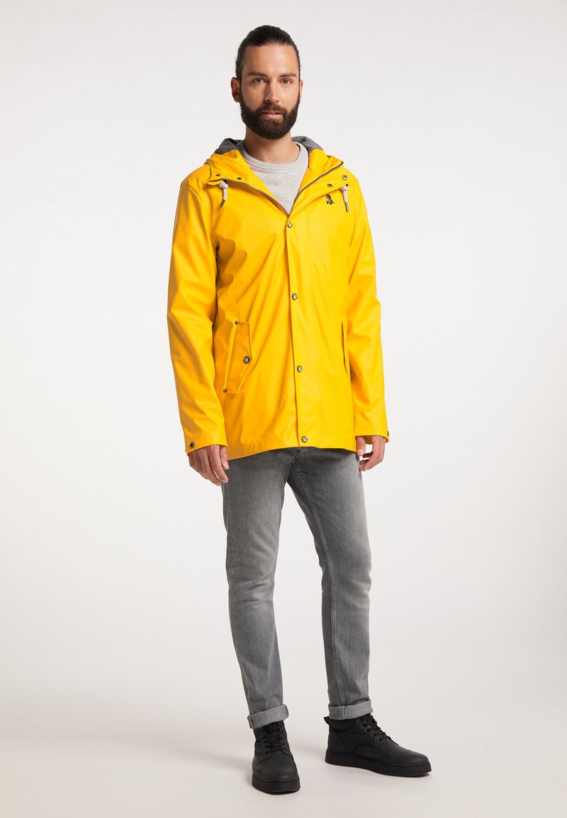 Gelbe wasserdichte Jacke mit kapuze, Reißverschluss und Druckknöpfen, zwei Fronttaschen und grauem Innenfutter. Getragen mit grauen Jeans und schwarzen Stiefeln.