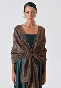 Shawl metallico marrone con una finitura testurizzata e scintillante, annodato al centro con un accento rotondo dorato. Indossato sopra un vestito verde.