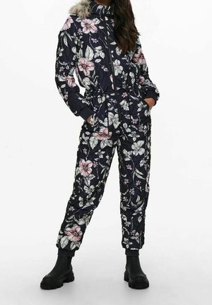 Kobieta w czarnym kwiecistym kombinezonie pikowanym z kapturem wykończonym futrem, z rękami w kieszeniach, w połączeniu z czarnymi botkami sięgającymi kostek.