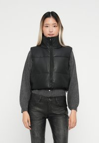 Melna pūkaina veste ar augsto apkakli, ar priekšējo rāvējslēdz�ēju, gludu tekstūru, izteiktām pildītām daļām, uzvilkta pār pelēku džemperi un melniem džinsiem.