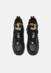Black high-top sneakers with textured finish, featuring contrasting yellow and white labels, round laces, and a rugged outsole design.