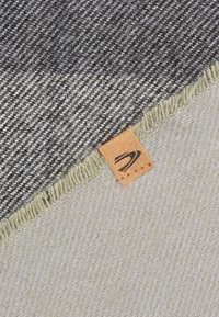 Uldblandings tæppe med et gråt og crème houndstooth mønster, frynsede kanter og et brunt lædermærke med et logo.