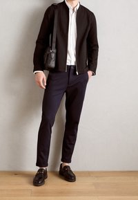 Homme portant une veste noire, une chemise blanche, un pantalon bleu marine, des mocassins en cuir noir, et portant un sac à épaule noir, debout contre un mur uni.