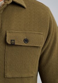 Zbliżenie na oliwkowozieloną, teksturowaną kieszeń koszuli z czarnym guzikiem i małą czarną metką PME Legend.