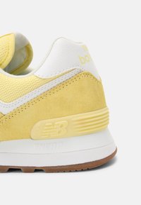 Close-up van de achterzijde van een gele en witte New Balance sneaker, gemaakt van suède en mesh materialen, met het NB-logo en een bruine buitenzool.