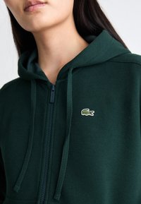 Grønn zip-up hettejakke med teksturert stoff, hette og snor. Har en liten brodert krokodillelogo på brystet.