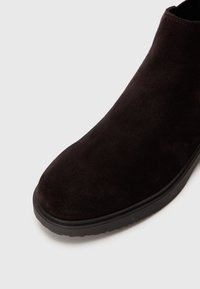 Bottine en daim noir à bout rond et semelle en caoutchouc noir sur fond blanc, inclinée pour montrer l'avant et le côté.