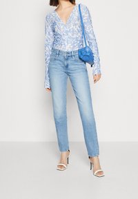 Hellblau gemusterte Langarmbluse mit tiefem V-Ausschnitt, kombiniert mit hellblauen geraden Jeans und weißen Riemchenabsätzen. Blauer Handtasche.