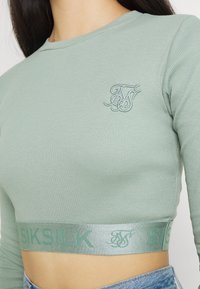 Kvinna som bär en mintgrön ribbad långärmad croptop med broderad logotyp och märkeselastisk nederkant, i kombination med ljusblå jeans.